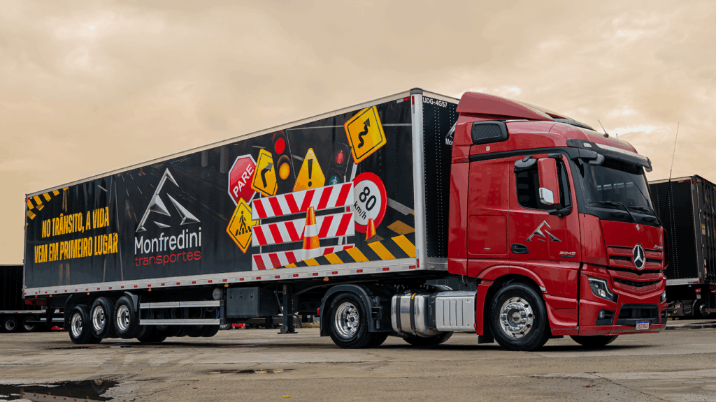 Caminhão vermelho da Monfredini Transportes com trailer ilustrado com placas de trânsito e mensagem de segurança viária